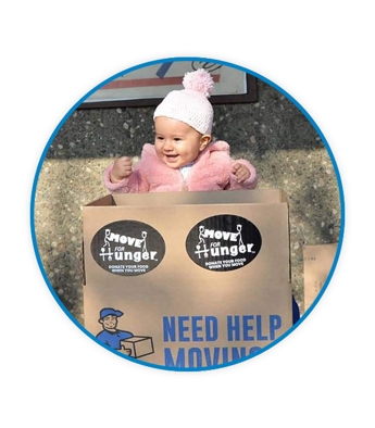 A baby in a pink jacket and snow beanie behind a Pete's Moving Services cardboard box