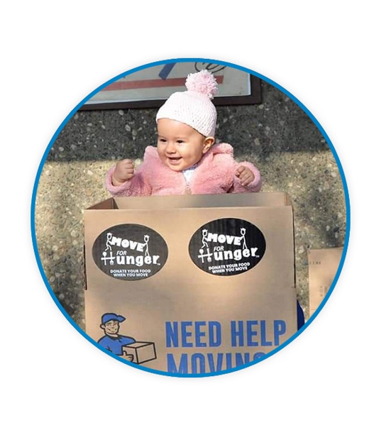 A baby in a pink jacket and snow beanie standing behind a Pete's Moving Service cardboard box